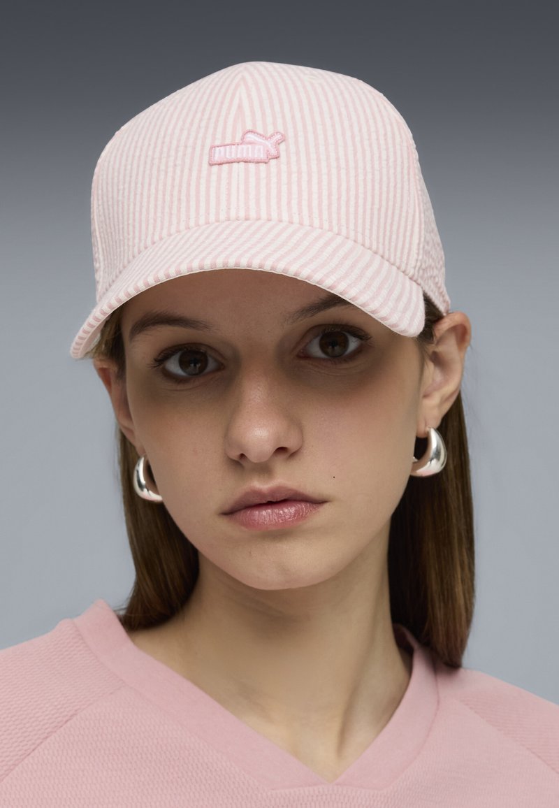 Jeune femme portant une casquette rayée rose pâle avec un petit logo brodé, des boucles d'oreilles anneaux argentées et un haut rose clair, sur un fond gris.