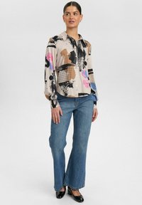 Mehrfarbig gemusterte Bluse mit langen Ärmeln, die abstrakte Designs in Pink, Schwarz und Blau zeigt, kombiniert mit weit geschnittenen blauen Jeans.