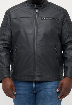 Homme portant une veste en cuir noire zippée avec des épaules renforcées et un jean bleu, debout les mains détendues sur les côtés.
