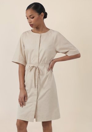 Woman wearing a beige knee-length dress with short sleeves and a waist tie, looking down with one hand on her hip.