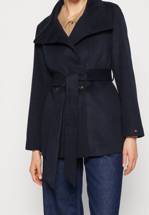 Mujer con un abrigo azul marino con cinturón sobre una camisa beige y jeans azules, de pie frente a un fondo blanco liso.