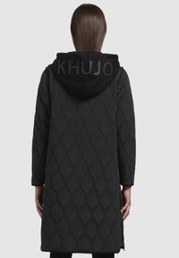 Cappotto lungo trapuntato nero con cappuccio e logo in rilievo, motivo a diamante, tessuto testurizzato e fessure laterali per una maggiore libertà di movimento.