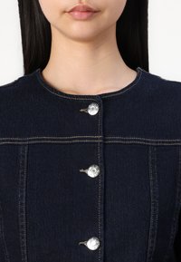 Mörk denimjacka med fem silverfärgade tryckknappar, kontrastsydda sömmar och rund halsringning. Har ett strukturerat tyg med en slät yta.