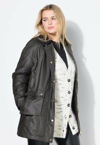 Giacca impermeabile nera con chiusura a zip, bottoni a pressione e tasche laterali. Abbinata a un cardigan a fantasia crema e pantaloni neri.