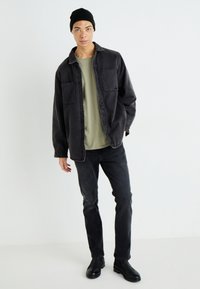 Chaqueta de mezclilla negra sobre una camiseta verde, combinada con jeans oscuros y botas negras. La chaqueta cuenta con dos bolsillos frontales y un corte relajado.