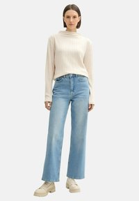 Pull en maille côtelée crème avec un col montant, associé à un jean bleu clair à jambes larges et des chaussures à plateforme beiges, se tenant devant un fond blanc.