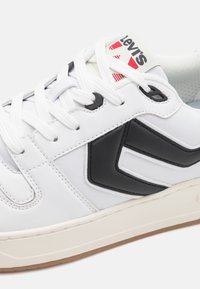 Baskets blanches Levi's avec motif chevron noir, lacets blancs et logo rouge sur la languette, avec une semelle beige et des détails cousus.