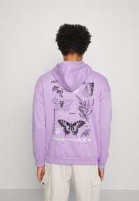 Felpa viola con cappuccio regolabile, caratterizzata da grafiche di farfalle nere e argento e testo sul retro. Texture morbida, design con vestibilità rilassata.