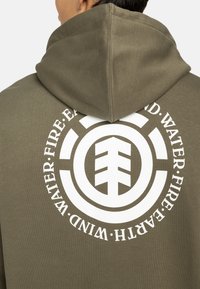 Sudadera con capucha en algodón verde olivo. Presenta un gran gráfico circular blanco con símbolos elementales en la parte posterior. Textura suave.