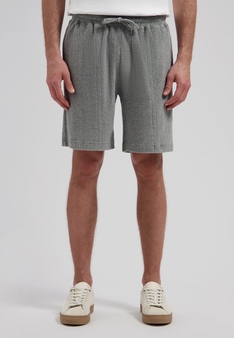 Homme portant un short gris matelassé à cordon de serrage et des baskets blanches, debout devant un fond uni.