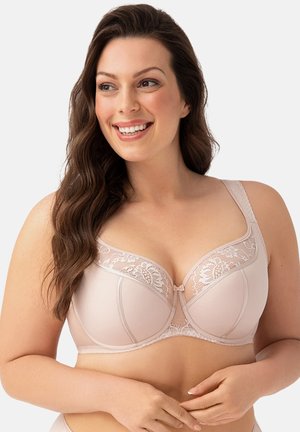 Smilende kvinde med langt brunt hår iført en lys rosa blonde- og mesh-bh, der kigger let til siden.