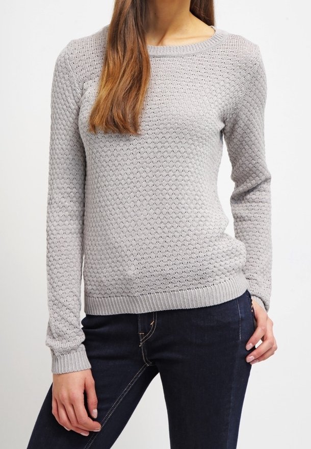 Femme portant un pull en maille texturé gris clair et un jean bleu foncé, avec une main dans la poche et de longs cheveux bruns sur l'épaule.