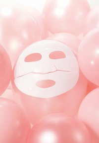 Des ballons en latex rose entourent un masque facial blanc avec des découpes pour les yeux et la bouche, créant un élément de design contrastant dans une texture douce.