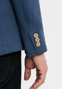 En mans hand hänger bredvid en marinblå jackärm med tre bruna knappar, som är påsatt en mörk denimjeans.