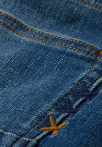 Tela de mezclilla azul con costuras naranjas en contraste y detalles decorativos de bordado multicolor. La textura parece ligeramente desvanecida.