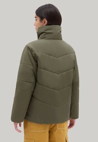 Chaqueta acolchada en verde oliva con un alto cuello y costuras en forma de chevron. Textura suave y diseño estructurado. Llevada sobre pantalones amarillos.