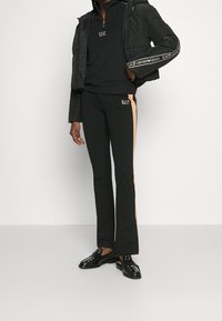 Černá péřová bunda se zipem a akcenty loga, v kombinaci s černými kalhotami, které mají kontrastní béžový boční pruh a branding. Černé boty doplňují outfit.