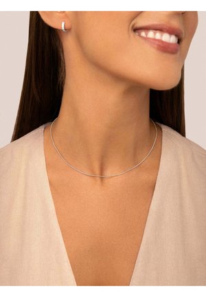 Mujer con cabello largo y oscuro, lleva un delgado collar de plata, un pequeño arete de plata en forma de aro, top beige con escote en V, sonriendo con los dientes visibles.