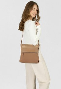 Bolso bandolera marrón con textura suave, que cuenta con un bolsillo frontal con cremallera y una correa ajustable, llevado por una persona con una blusa blanca.