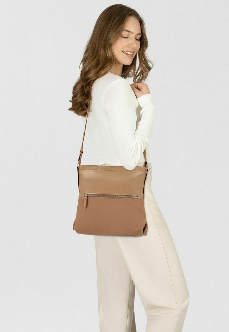 Bolso bandolera marrón con textura suave, que cuenta con un bolsillo frontal con cremallera y una correa ajustable, llevado por una persona con una blusa blanca.