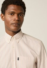 Chemise boutonnée rayée rose clair et blanche avec un col pointu, poche avant et un petit patch de logo sur la poche. Tissu texturé.