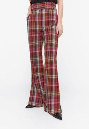 Femme portant un pantalon à carreaux taille haute, jambes larges, dans des tons rouges, roses et gris, avec des chaussures noires à bout pointu, debout les mains dans les poches.
