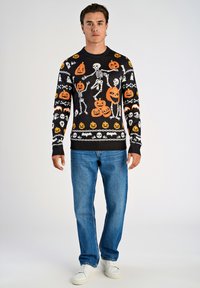 Czarny sweter z dzianiny z wzorami czaszki i dyni w białym i pomarańczowym kolorze, okrągły dekolt i ściągacze. Połączony z niebieskimi dżinsami.