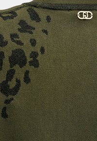 Tessuto lavorato a maglia verde oliva con macchie leopardate nere e una piccola spilla in metallo dorato nella zona del colletto.
