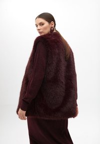Bordový faux fur vest s texturovaným povrchem, s volným střihem a dlouhými rukávy, v kombinaci s ladícím šaty a výraznými náušnicemi.
