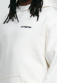 Sweat à capuche blanc avec une poche avant, une capuche à cordon ajustable et un petit logo "lowlights" noir sur la poitrine. Texture de tissu douce.