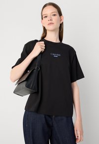 Black cotton T-shirt with short sleeves, featuring a blue "Calvin Klein Jeans" logo. Model holds a small black handbag with a flap.