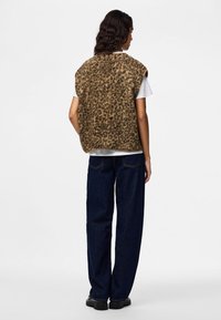 Persona ar gariem tumšiem matiem valkā bez piedurknēm brūnu rakstainu vestes virs baltas īsroku krekla, tumši zilas džinsas un melnus apavus, stāv ar muguru uz skatītāju.