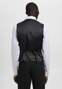 Schwarze Satinweste mit glatter Textur, figurbetontem Schnitt, verstellbarem Rückengurt und kombiniert mit einem weißen Langarmhemd.