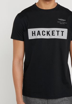 Czarna bawełniana koszulka z szarym prostokątnym logo z napisem "HACKETT" i małym logo Aston Martin Racing na klatce piersiowej.