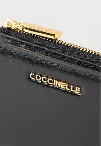 Musta nahkainen lompakko kiiltävällä pinnalla, jossa on kultainen vetoketju ja metalliosat sekä kultainen kohopainettu Coccinelle-logo edessä.