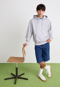 Un sweat à capuche gris avec une poche avant, associé à un short en denim bleu. Se tenant à côté d'une chaise en bois sur un sol en moquette verte.