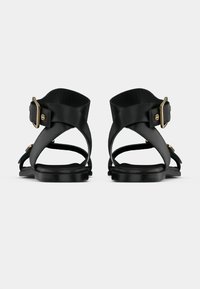 Ζευγάρι μαύρων δερμάτινων πέδιλων με επίπεδη σόλα και λουράκια στον αστράγαλο με αγκράφες σε χρυσό τόνο, εμφανιζόμενα από πίσω σε απλό φόντο.