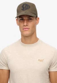 Casquette de baseball vert olive avec logo brodé, associée à un t-shirt en tricot beige présentant des rayures discrètes et un petit nom de marque brodé.