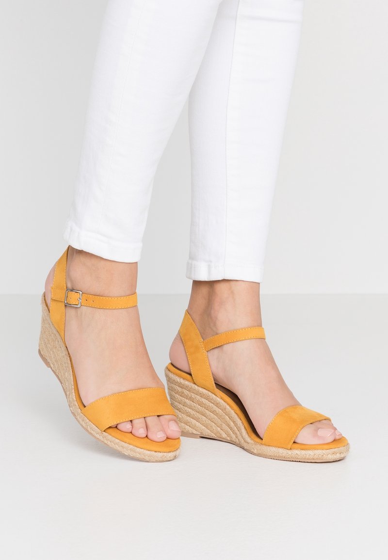 Pieds portant des sandales compensées jaunes avec brides à la cheville et pantalon blanc sur fond blanc.