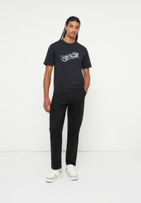 T-shirt grafica nera con design bianco, abbinata a pantaloni cargo neri e sneakers beige con accenti bianchi. Il modello è in piedi contro uno sfondo bianco.