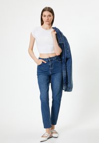 T-shirt blanc court, jeans en denim bleu taille haute avec une coupe droite, et une veste en denim bleu drapée sur l'épaule. Chaussures plates blanches.