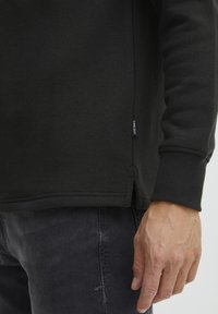 Svart hettegenser med ribbestrikkede mansjetter og nederkant, sidespalter, og et lite merkelapp med "PROJECT" på den nedre sømmen. Strukturert stoff, avslappet passform.