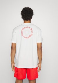 Valkoinen t-paita lyhyillä hihoilla, jossa on punainen teksti ja geometrinen muotoilu selässä. Yhdistetty punaisiin verkkoshortseihin.