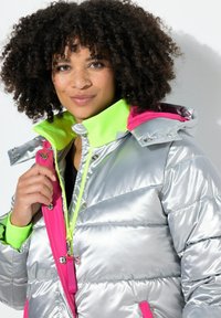 Veste puffer métallique argentée avec un col haut en néon vert et des accents roses ; elle dispose d'une fermeture éclair à l'avant et de boutons-pression.