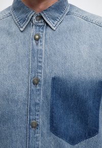 Gros plan d'une chemise en denim bleu clair avec une poche poitrine bleu foncé et des boutons-pression métalliques sur le devant.