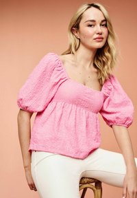 Blouse rose texturée avec de courtes manches bouffantes et un décolleté carré, associée à un pantalon blanc ajusté. Les détails incluent un tissu froncé.