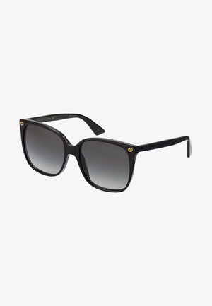 Gafas de sol negras de gran tamaño con lentes gradientes, montura de plástico grueso y acentos dorados del logo en las varillas. Forma angular y acabado pulido.