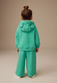 Turquoise hoodie with a rounded hood featuring ear accents, paired with wide-leg trousers in the same color. Soft, textured fabric.