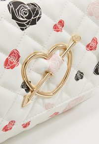 Prošívaná bílá taška se vzorem červených, černých a růžových růží, se zlatým srdcovým uzávěrem a detaily šípu.