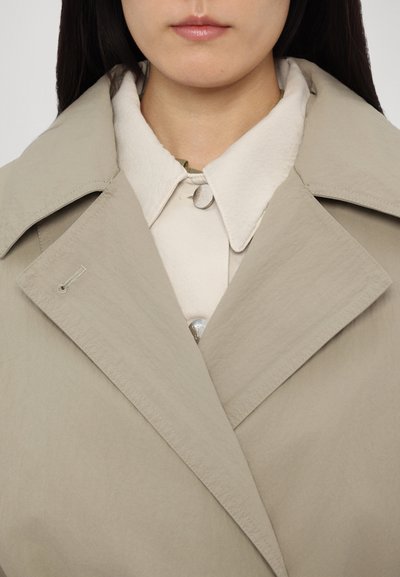 Light beige trench coat with a sharp collar, featuring a buttoned cream shirt underneath and subtle texture, closely cropped at the chest.